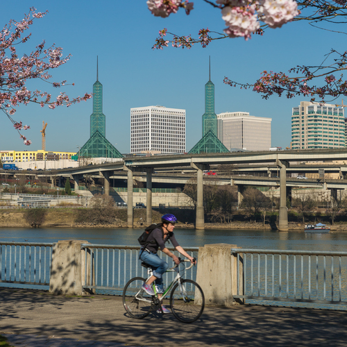 A University of Portland student riding their bicycle along a paved river front path with the Willamette river and city skyline in the background.