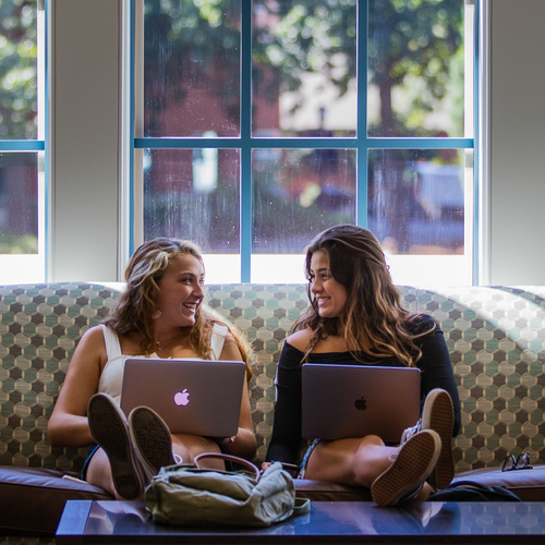 Two students holding laptop computers sit on a plush sofa with their feet propped up on a table and smile at one another in Dundon Berchtold Hall at the University of Portland.