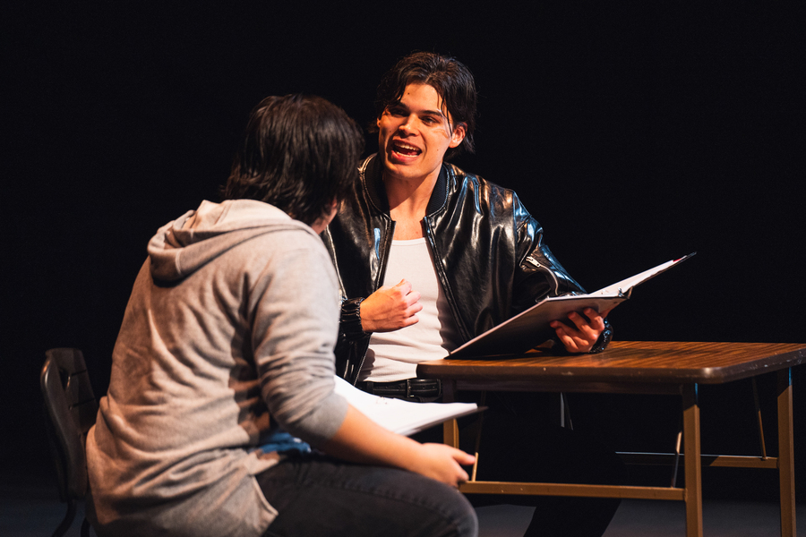 Two University of Portland theater students sit at a table on stage while acting out a scene from the production of TBA.