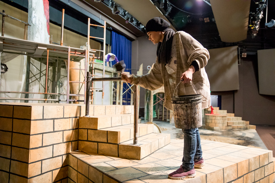 A University of Portland theater student wearing and scarf and beret stands in a partially built stage set, intently painting a scenery element.