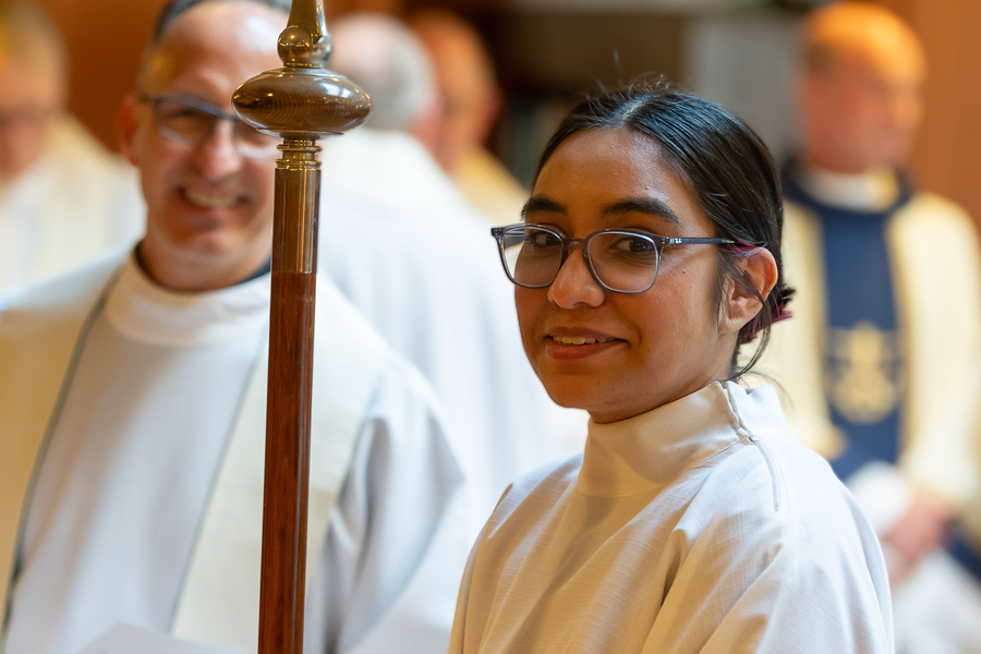 A campus ministry student smiles at the camera while acting as a server during mass in the Chapel of Christ the teacher at the University of Portland, a priest smiles in the blurred background.