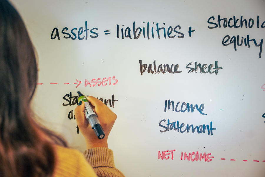 The back of a student's head and hand are visible at the edge of the image as they write accounting notes and terms on a whiteboard.