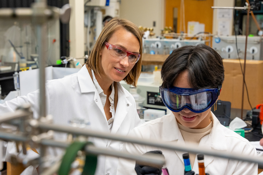 A professor watches over a student, both dressed in lab coats and goggles, working with chemicals in a laboratory at the University of Portland.