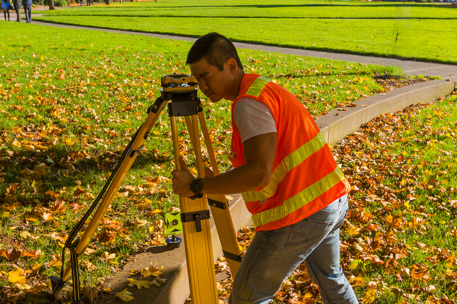 A UP civil engineering student peers into a yellow surveyors tripod on grass scattered with autumn leaves.