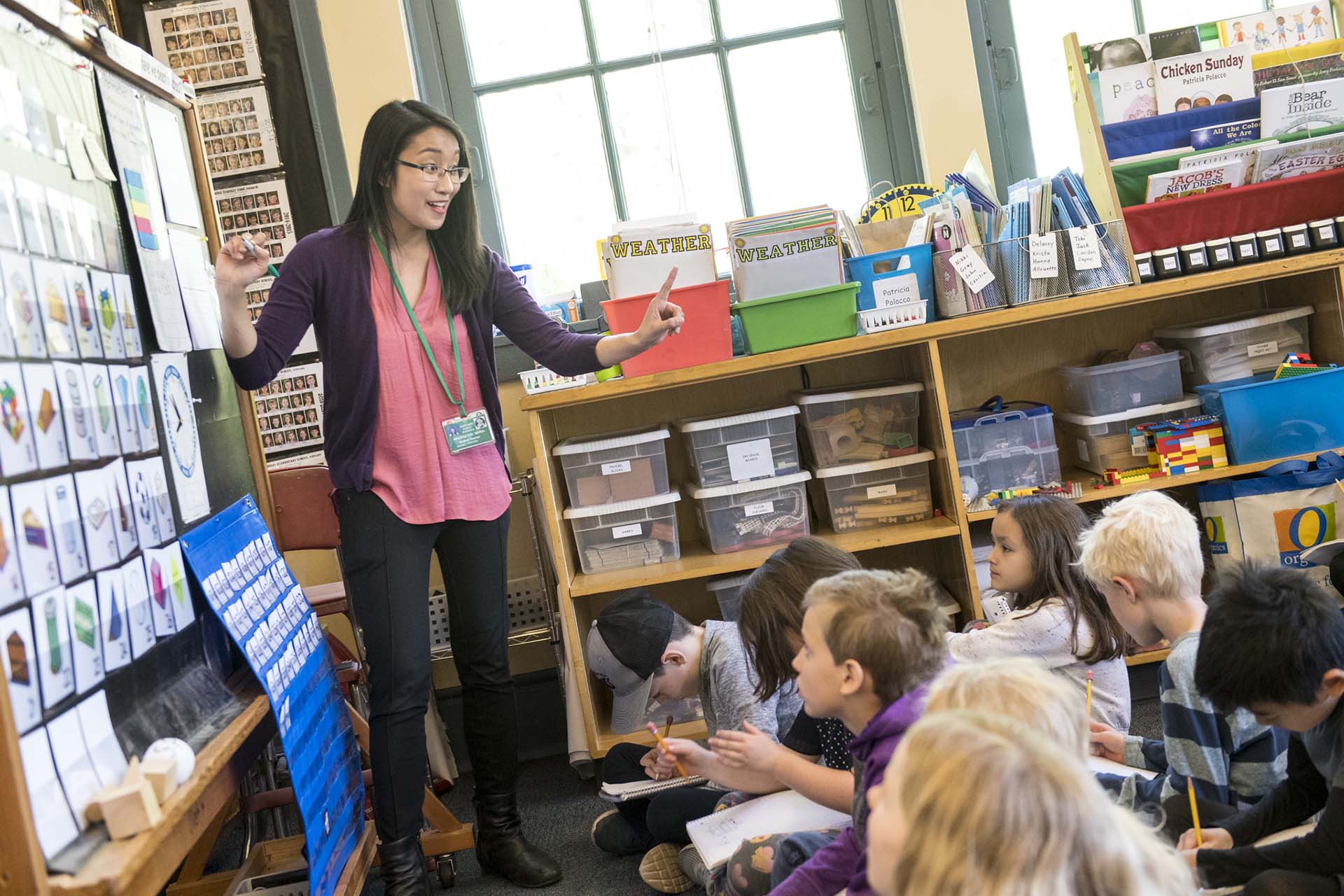A University of Portland education student stands in front of an elementary class of children sitting on the floor and points to letters and numbers on the board