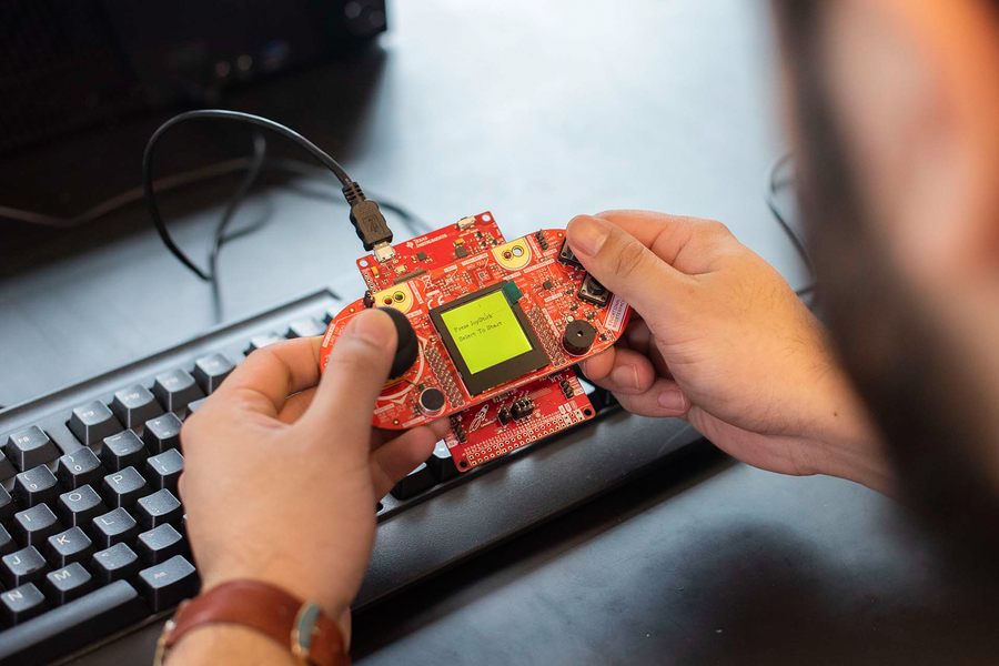 A close-up of a persons hands manipulating a circuit board, with part of the desk, keyboard, and monitor stand visible in the background.