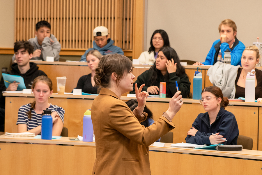 A University of Portland English professor stands at the front of a tiered classroom during a lecture facing towards the students, who listen intently.