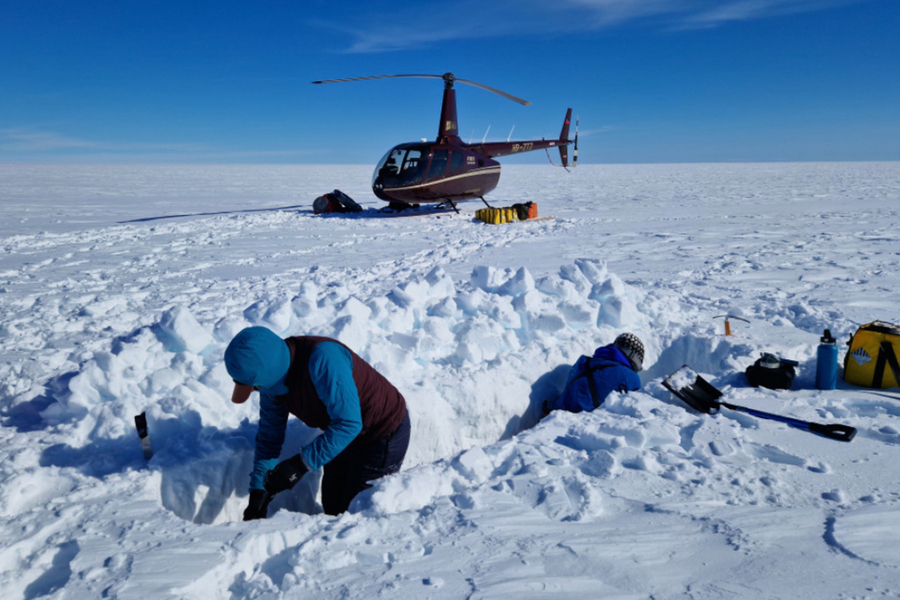 A helicopter is parked on a vast, snowy landscape under a clear blue sky. A UP environmental science student is in the foreground digging in deep snow surrounded by scientific instruments. 