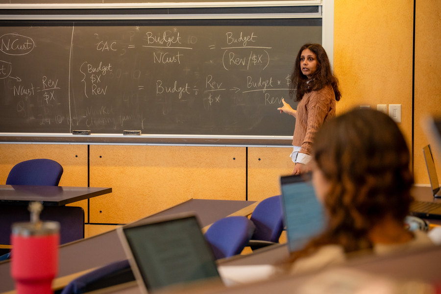 A UP Finance professor stands in front of a chalkboard displaying handwritten budget formulas and financial terms; partial view of a student's face appears at the image edge.