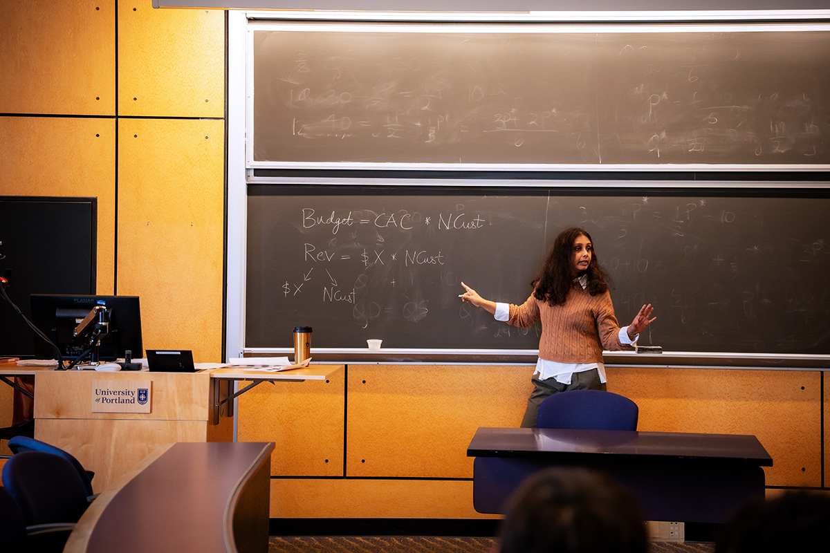 A University of Portland Business Finance professor points to a blackboard with equations during lecture while facing and speaking to students