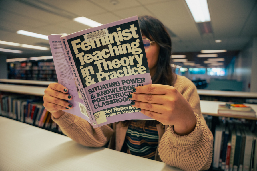 A student is seated at a table, their face obscured by the feminist theory book that they are reading in the Clark Library at the University of Portland.