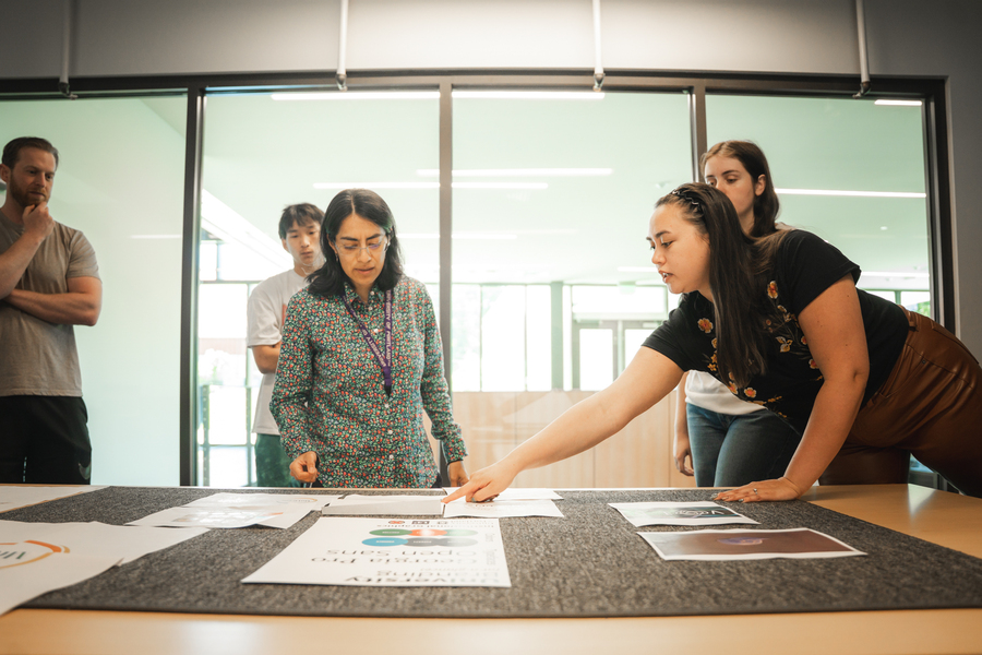 Four UP students and their professor are engaged in thoughtful dialogue over project papers laid out on a table in the Innovation Think Tank classroom.
