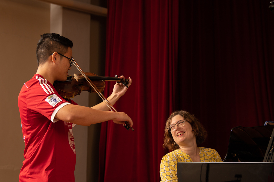 A music student stands on stage playing their violin while their smiling professor accompanies them on a piano at the University of Portland.