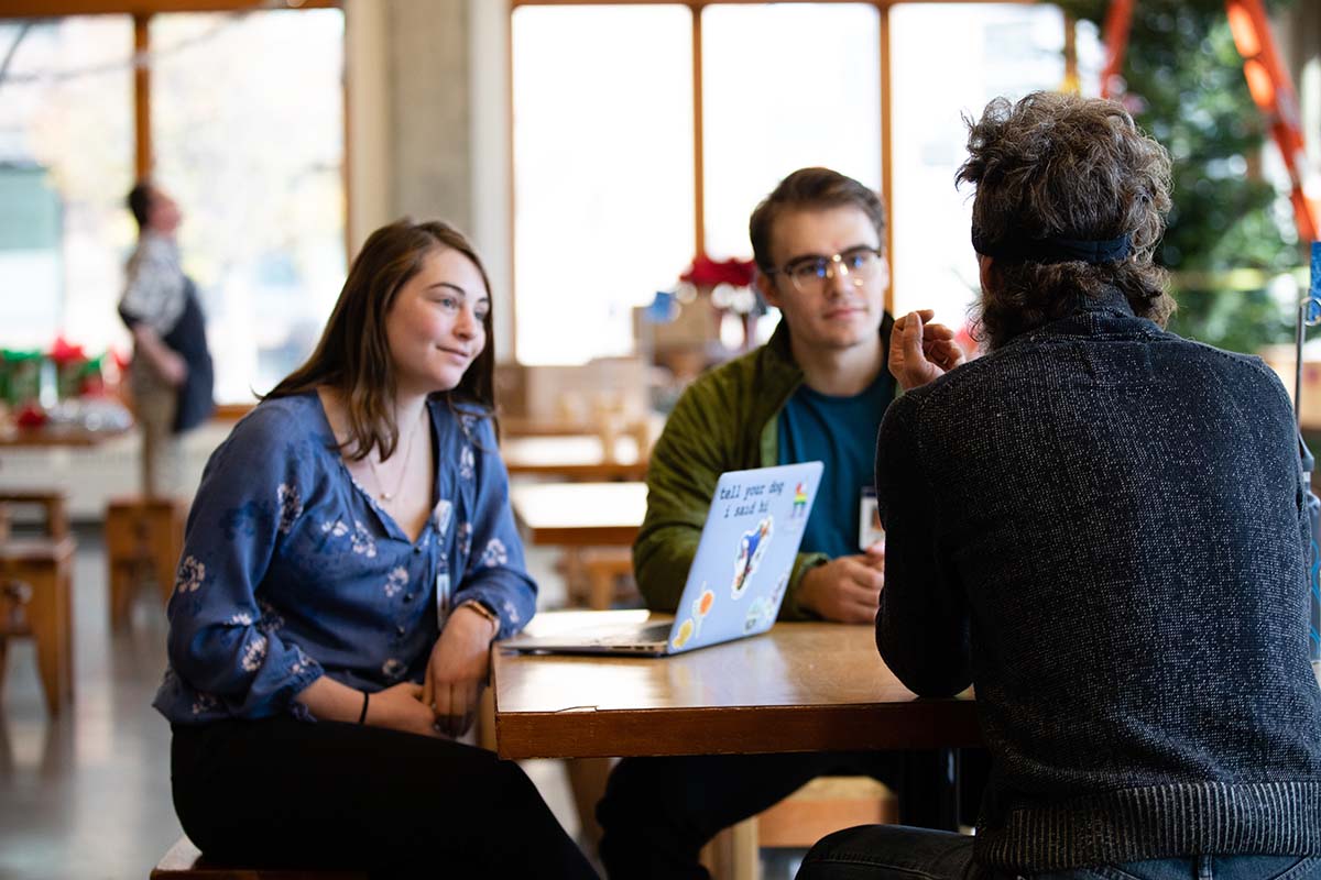 Two University of Portland students sit at a table with a  member of the Blanchet House non-profit with Christmas decorations being hung in the background