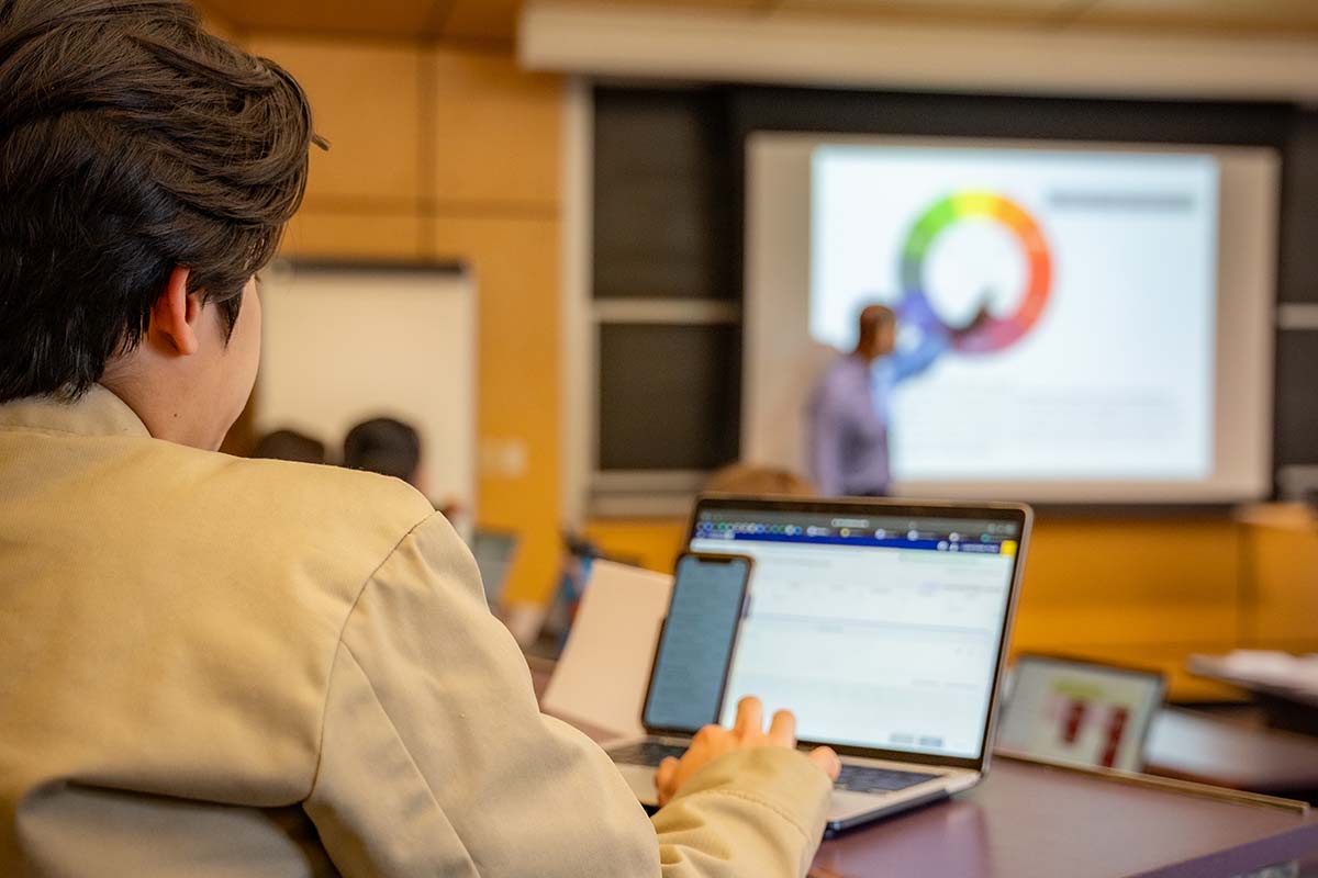 A University of Portland student takes notes on a laptop while professor points to a colorful pie chart on the projector screen in the background during a strategic management lecture