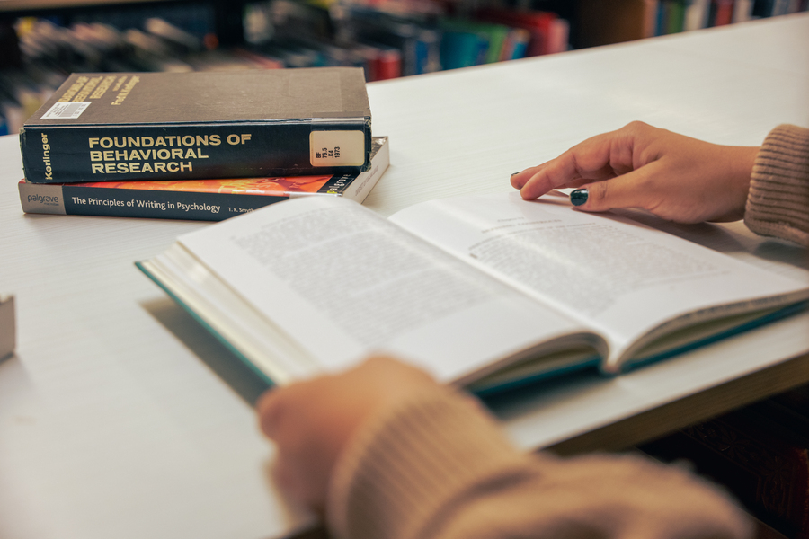 Close-up of student hands on an open book with blurred text; a stack of psychology text books is partially visible in the background.
