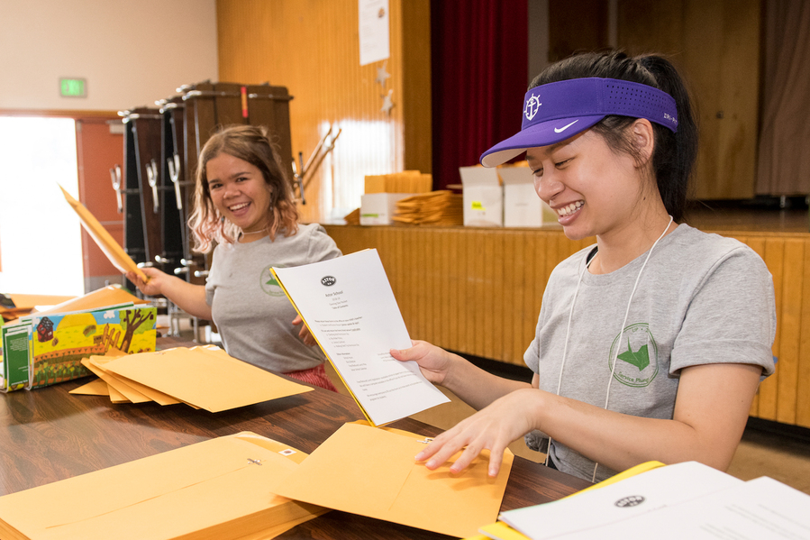 A smiling UP student in a gray shirt sits indoors next to a stack of paperwork. The image is bright and cheerful, with papers visible in the foreground.