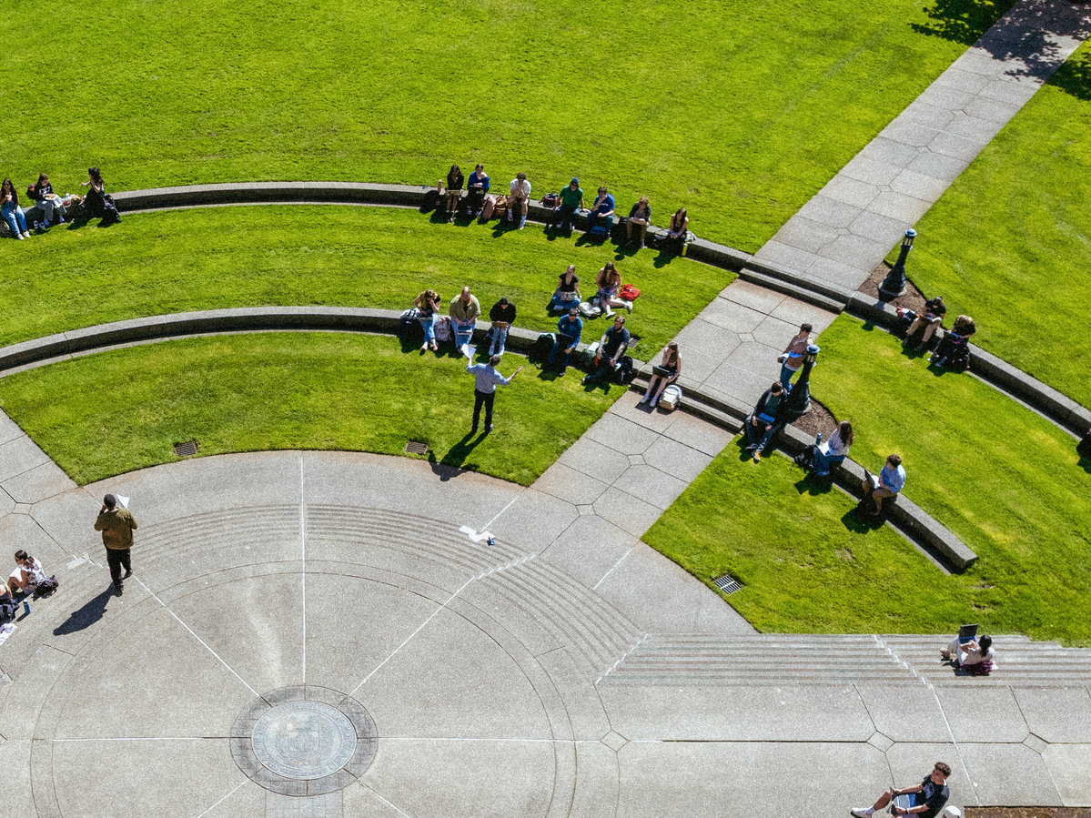 An aerial view of students sitting in a large semicircle on the grass and on concrete steps for an outdoor class.