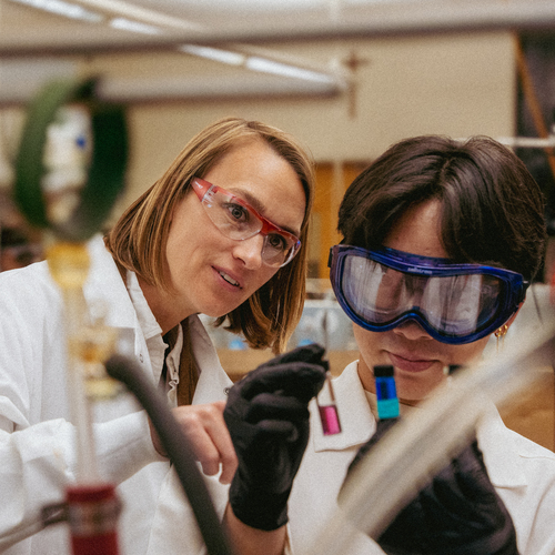 A professor instructs a student holding two small vials in a science lab, both wearing safety goggles and lab coats.