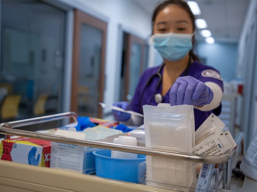 A nurse reaches a gloved hand into a supply cart in a healthcare setting.