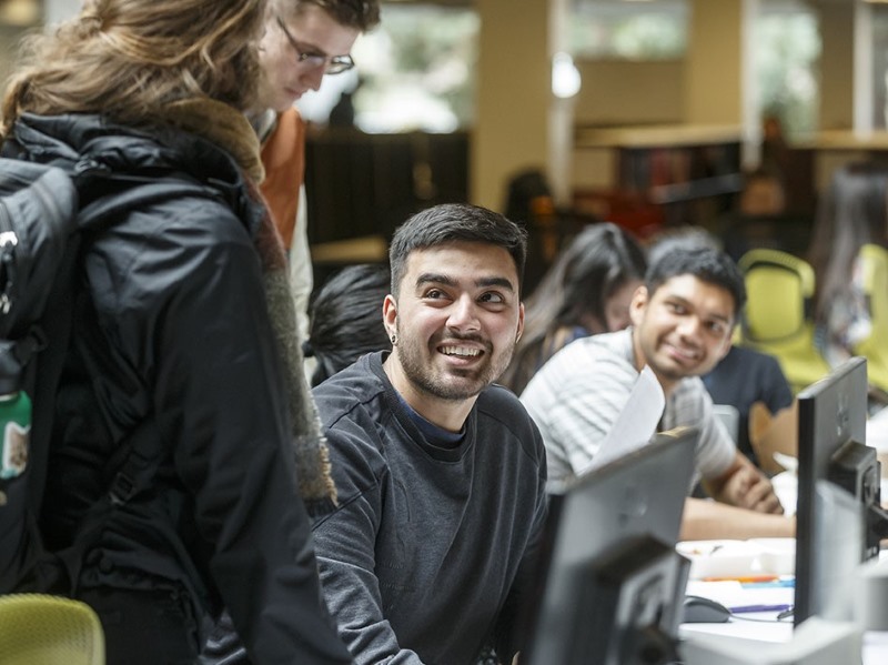 Students sitting at a row of computers in the Clark Library, smiling up at a standing student