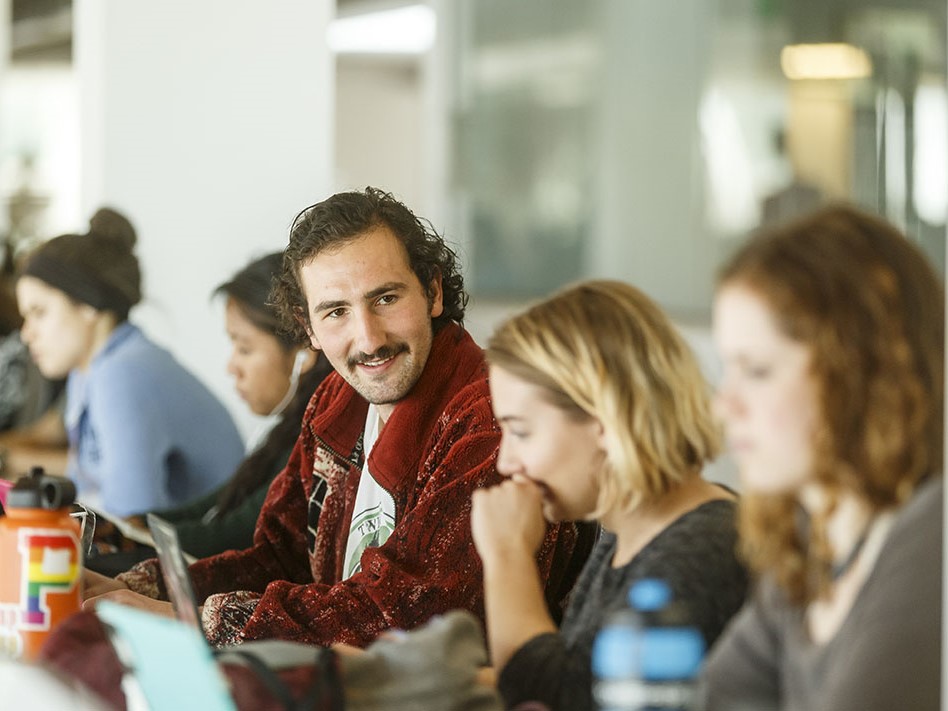 A student with a mustache looks over, smiling at his peers in a classroom