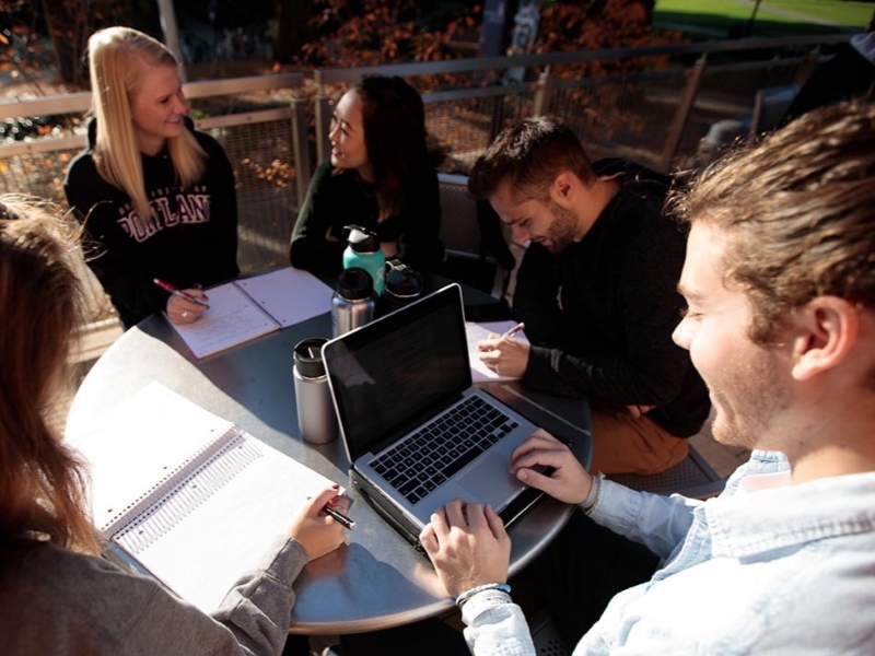 A group of friends sit at a table outside, one student looking at their laptop screen