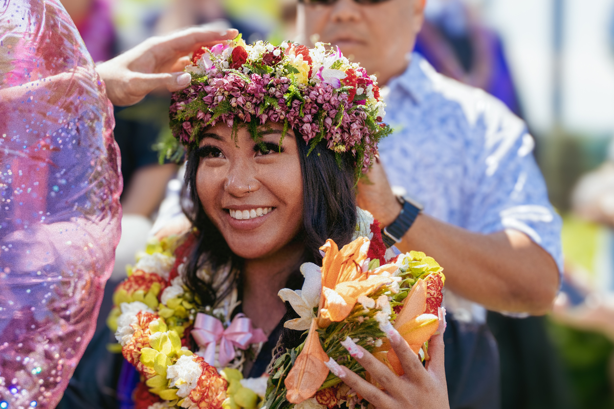 A graduating student smiles at commencement while a crown of flowers is placed on their head.