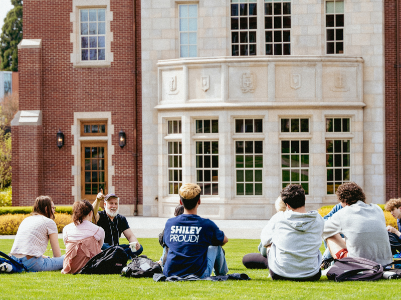 A small group of students and their professor sitting on the lawn in front of a campus building having class outside.