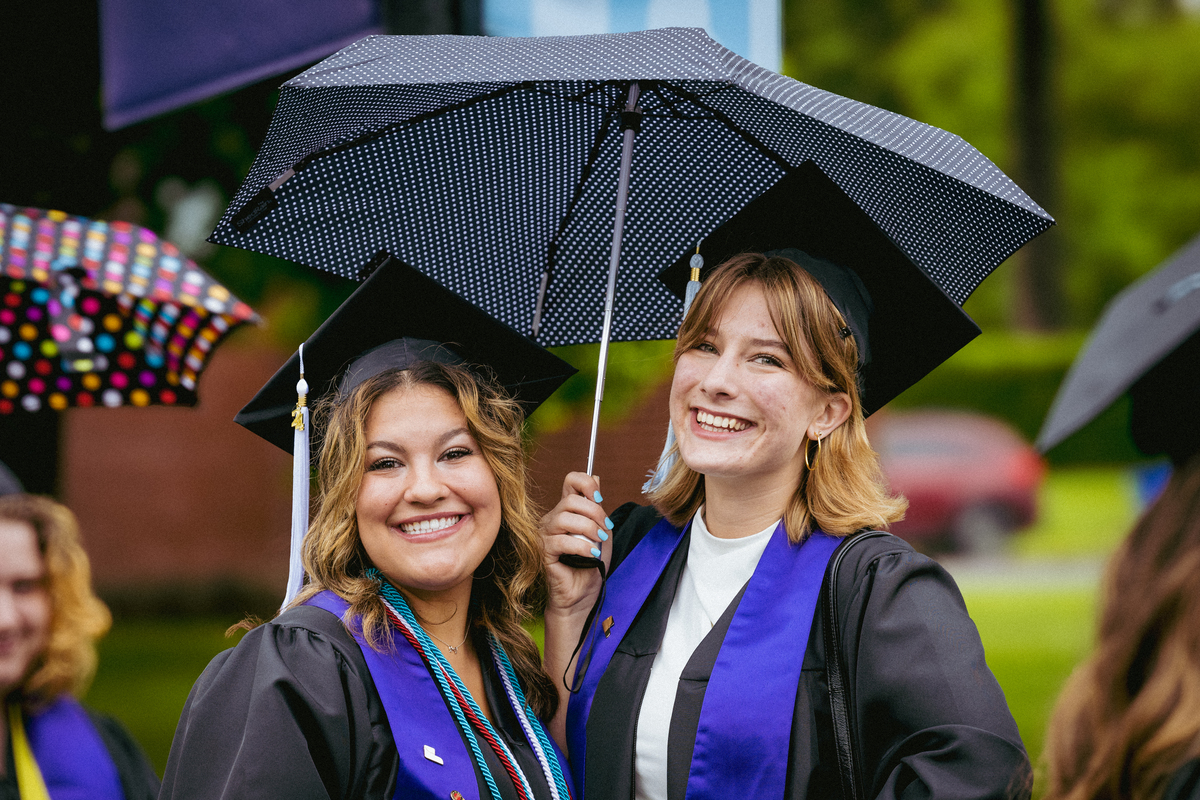 Two University of Portland graduates wearing their caps and gowns smile under a polka dot umbrella on graduation day.