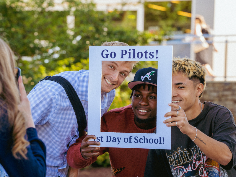 Three University of Portland students smile and hold a white photo frame that says "Go Pilots! 1st Day of School!"