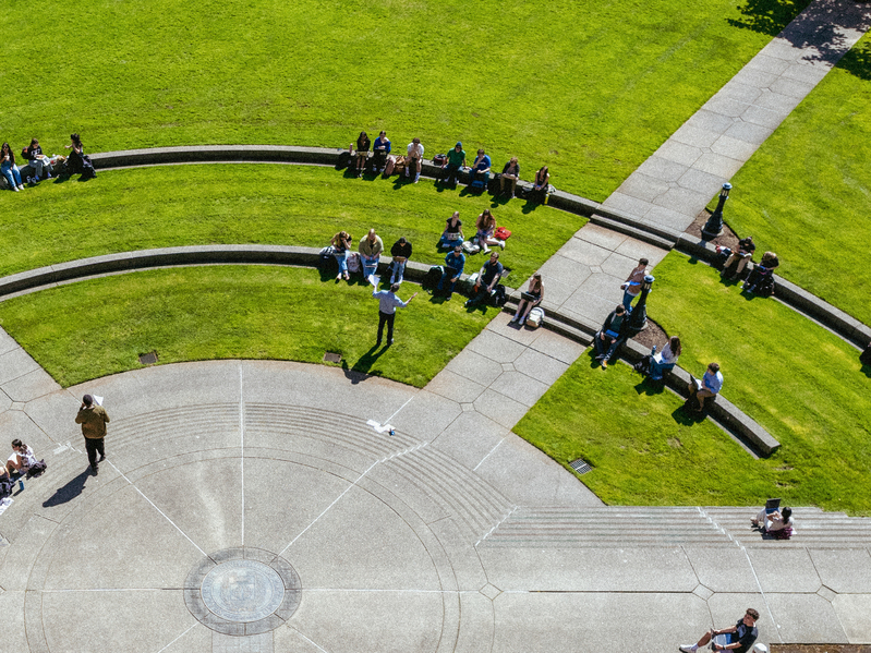 An aerial view of students sitting in a large semicircle on the grass and on concrete steps for an outdoor class.