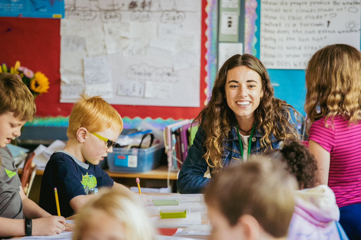 An elementary education major smiles while sitting at a table with students in a classroom at Duniway Elementary School.