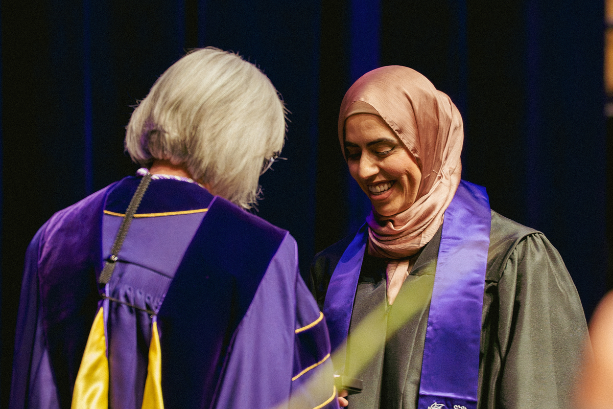 A graduating nursing student wearing a hijab and graduation gown receives a pin from a professor at a pinning ceremony.