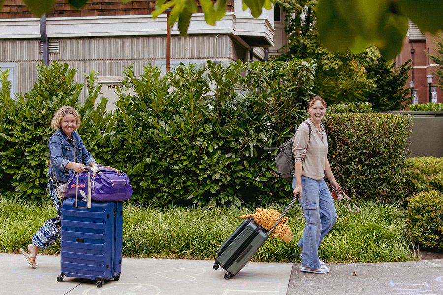 A parent and student with luggage and backpacks walk on a sidewalk on campus, smiling during move-in day.