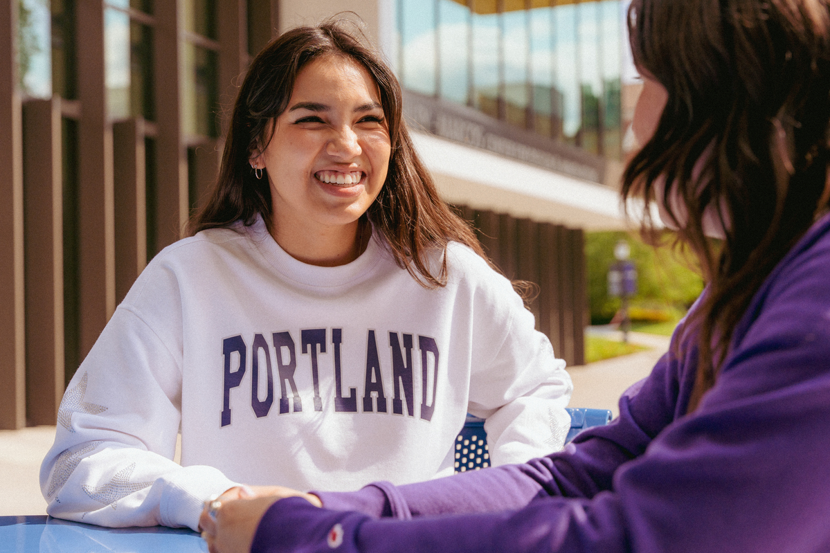 A student in a "Portland" sweatshirt sits at an outdoor table, smiling and talking to their admissions counselor.