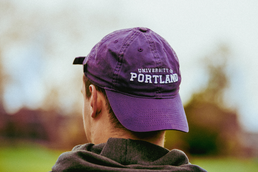 A behind view of a University of Portland student wearing a purple University of Portland baseball cap backwards. 