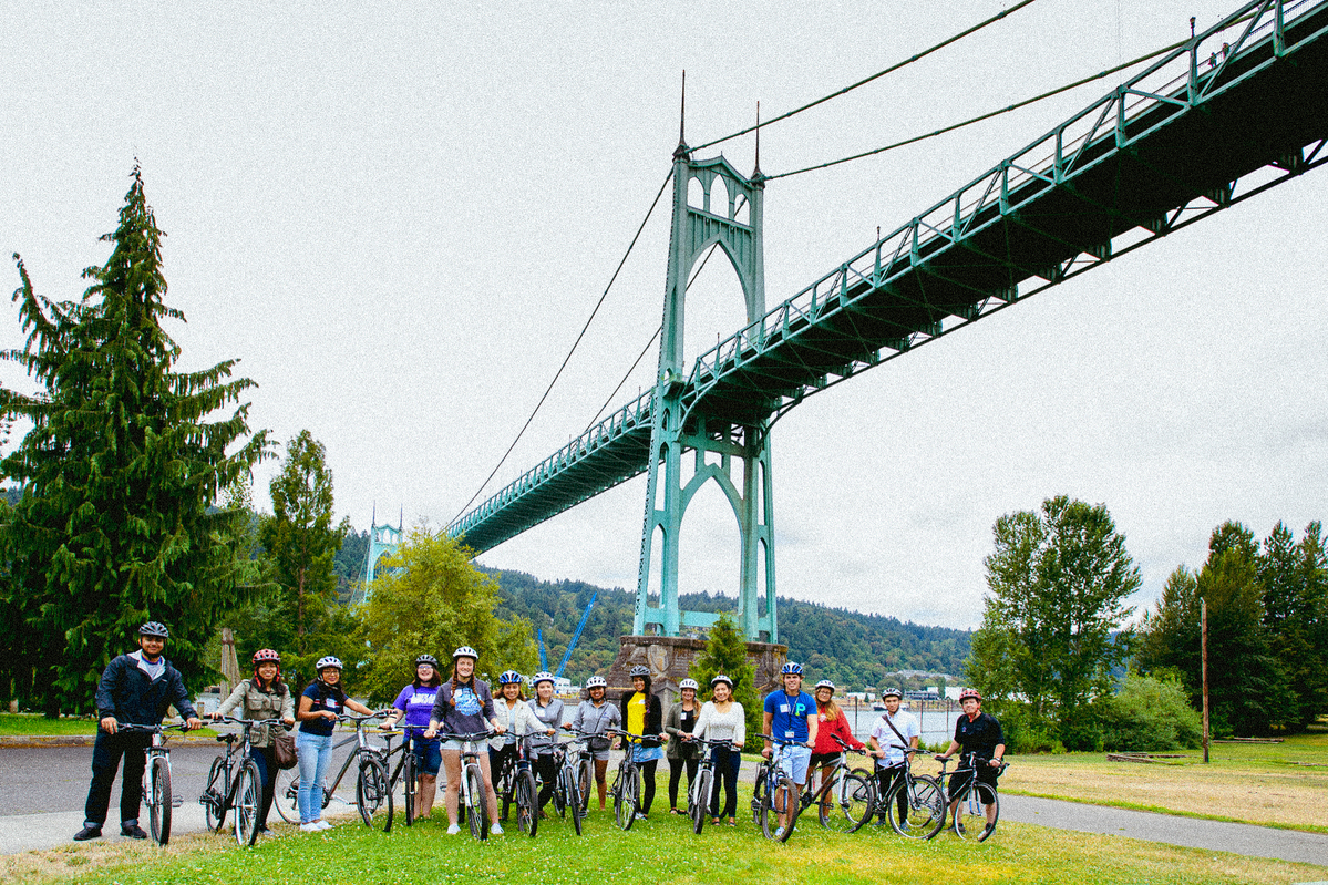 A group of students wearing helmets stand with their bicycles in Pier Park, with the St. Johns Bridge in the background.
