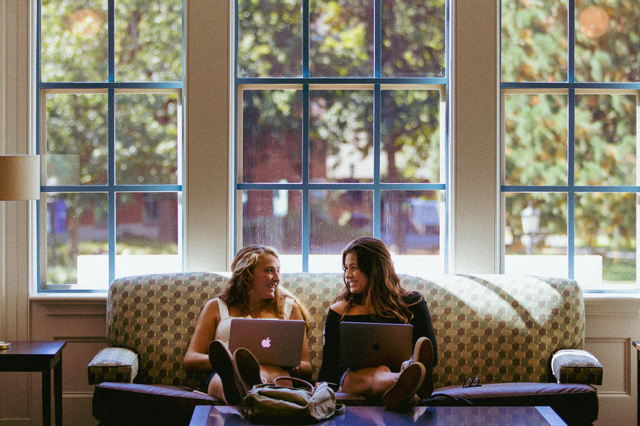 Two students sit on a couch in Dundon Berchtold Hall with their laptops open, talking and smiling.