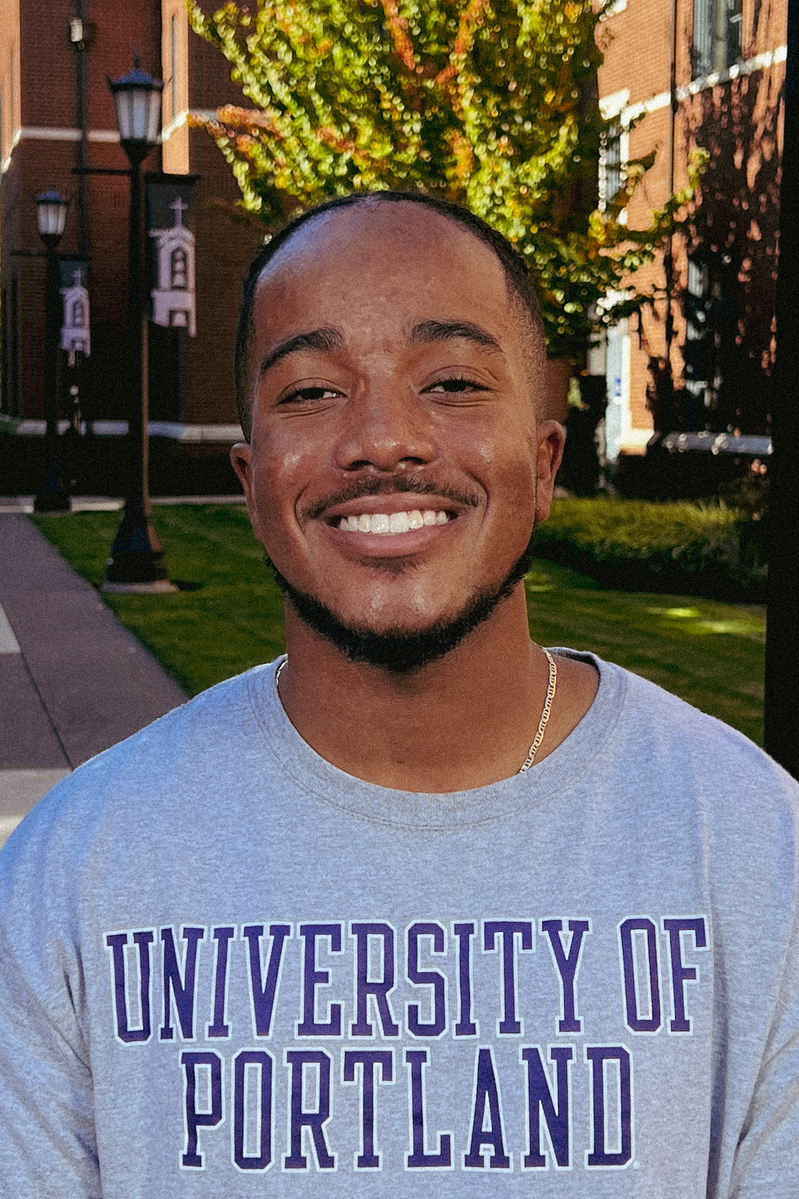 Darius Newman smiles for a headshot in front of campus buildings, wearing a gray and purple University of Portland shirt.