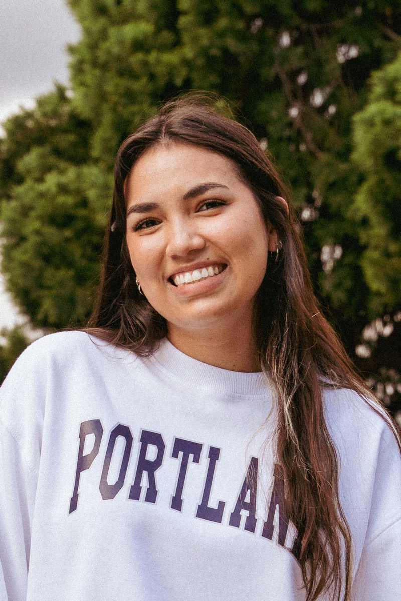 Laya Duguid smiles for an outdoor headshot, wearing a white sweatshirt that says "Portland" in purple.