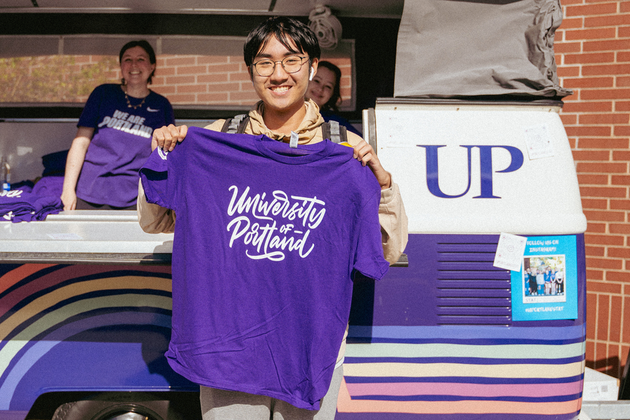 A student smiles and holds a purple t-shirt that says "University of Portland" in front of the Pilot Van.