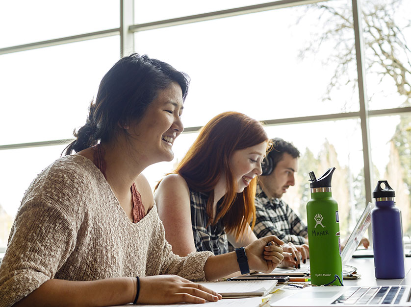 Three students sit in a common area working on their laptops, smiling