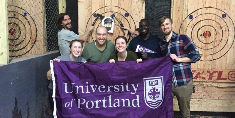 Alumni holding a UP flag and axes in an axe throwing alley an axe throwing 
