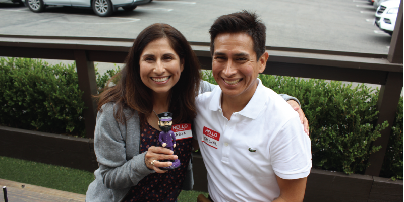 A couple posing at a picnic table with a bobble-head of Wally the Pilot. 