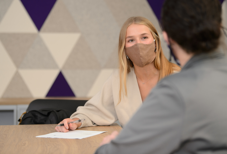 A student and staff member sit at a conference table. They are wearing face masks. 
