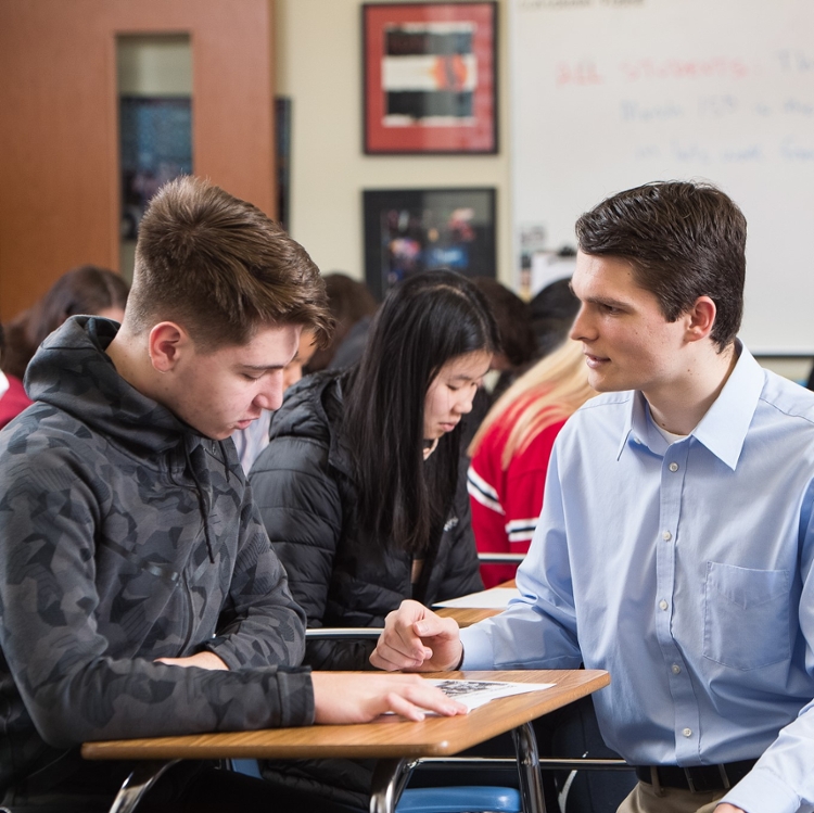 male student teacher kneeling by male youth in desk discussing paperwork.