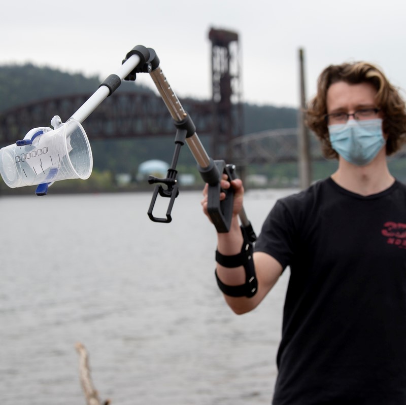 masked student with trash collection arm device holding clear plastic cup near river