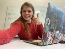 student sitting at desk with laptop and thumbs up to camera