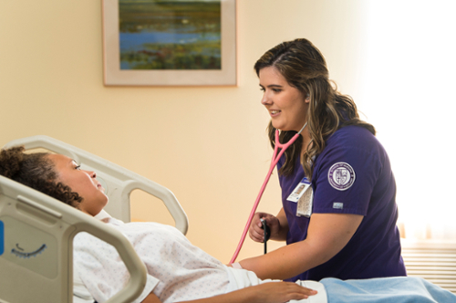 UP nurse in purple scrubs tends to patient in hospital bed